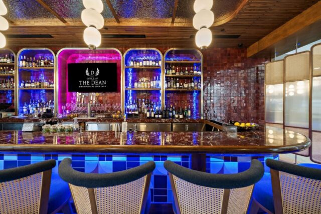 A photo of the bar at the Dean. The bars mid century style lighting hangs down from a swirling silver metal ceiling reflecting back the bright lights and giving the bar a playful atmosphere. The backwall of the bar are deep red tiles in a variety of tones that give off a warm and cozy atmosphere. The bar is curving and allows the patrons to sit in comfortable blue velvet chairs as they watch the latest game.